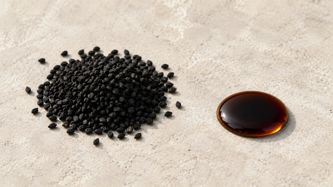 Matte black Nigella sativa seeds beside a single drop of deep amber black seed oil on a pale stone surface in soft daylight