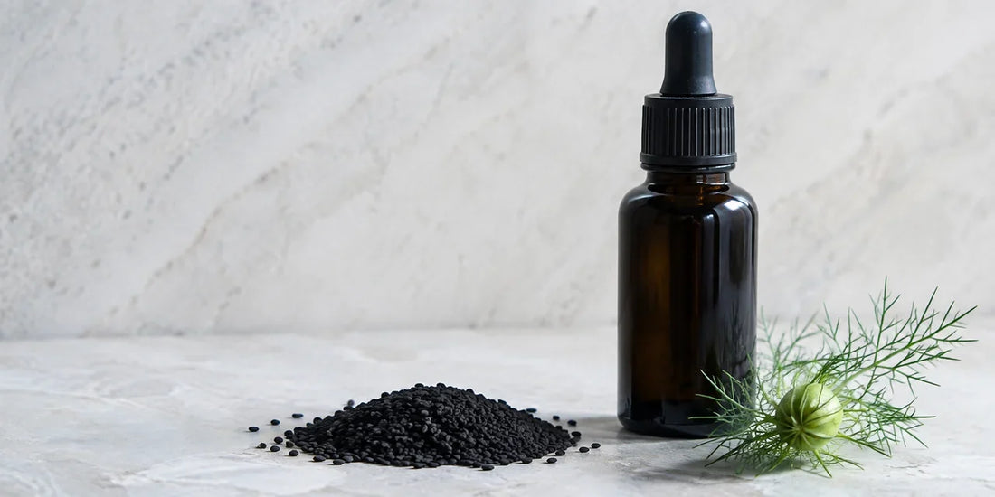 Matte black Nigella sativa seeds and a dark glass dropper bottle of deep amber oil on pale stone