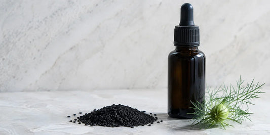 Matte black Nigella sativa seeds and a dark glass dropper bottle of deep amber oil on pale stone