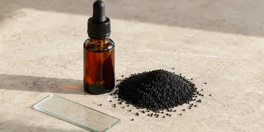 Dark glass dropper bottle of black seed oil beside a heap of whole black cumin seeds and a plain glass slide