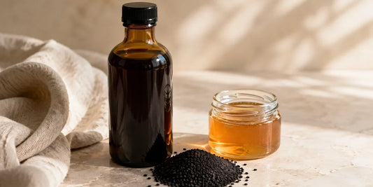 Dark glass black seed oil bottle beside matte black seeds and a honey jar on pale stone