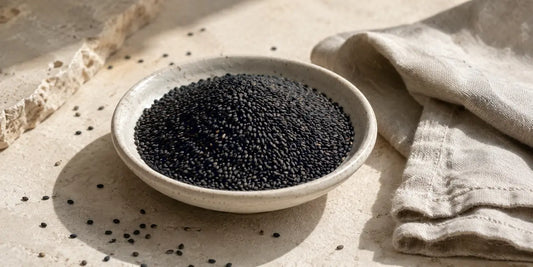 Shallow pale ceramic dish heaped with glossy black sesame seeds beside folded linen on a limestone surface