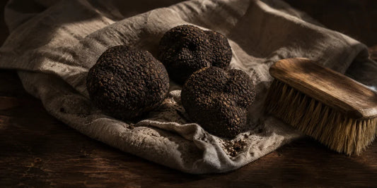 Three whole black truffles on crumpled grey linen beside a bristle brush on a dark wooden table