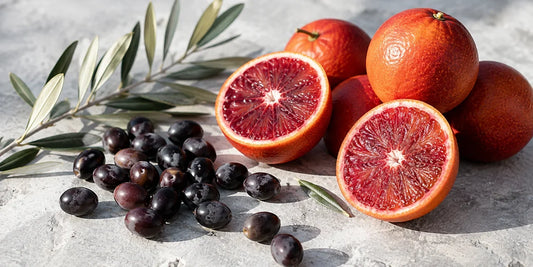 Halved blood oranges showing crimson flesh beside dark olives and leaves on pale grey stone
