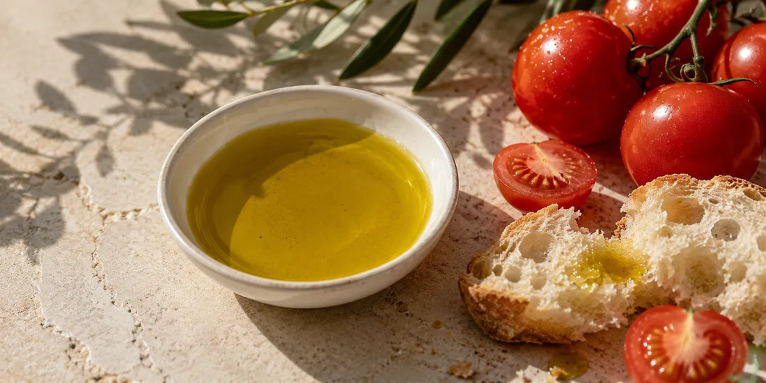 Shallow white bowl of rich golden-green olive oil beside ripe tomatoes and torn bread on pale stone