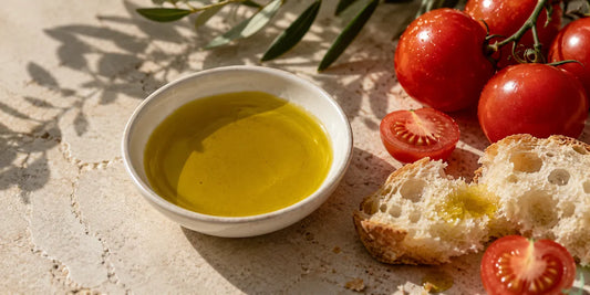 Shallow white bowl of rich golden-green olive oil beside ripe tomatoes and torn bread on pale stone