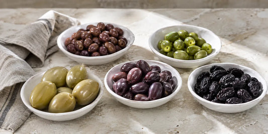 Shallow white bowls of assorted olives, large green, small dark and purple-black, on a pale stone counter