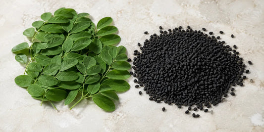 Fresh green moringa leaves beside a heap of matte black Nigella sativa seeds on pale stone in daylight