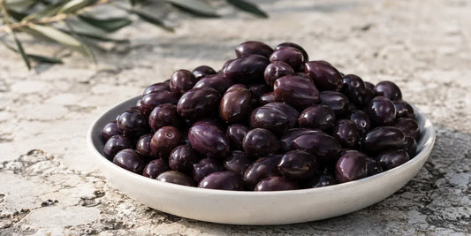 Very small dark purple-black olives glossy with brine heaped in a shallow white dish on pale stone