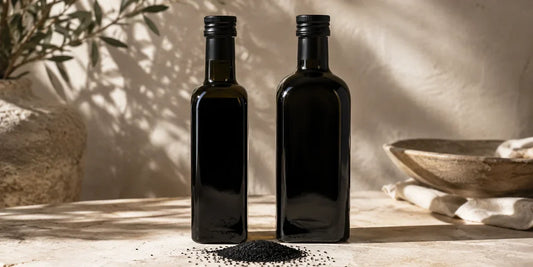 Two dark glass black seed oil bottles side by side on a pale stone surface with Nigella seeds in warm directional light