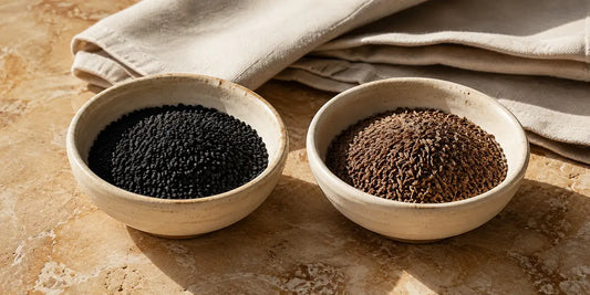 Two small pale bowls of different dark seeds side by side on a warm stone surface in soft light