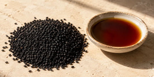 Matte black Nigella sativa seeds beside a shallow ceramic dish of deep amber oil on pale stone