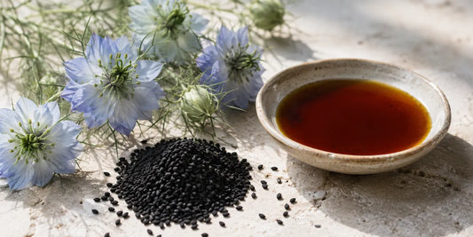Pale blue Nigella sativa flowers beside black seeds and a dish of deep amber oil on limestone