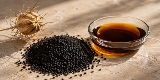 Heap of matte black Nigella sativa seeds beside glass dish of deep bronze oil on pale stone