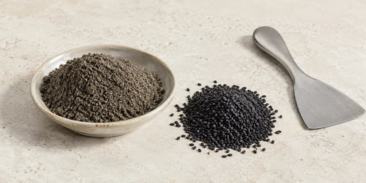 Shallow dish of fine dark seed powder beside a heap of matte black sesame seeds on pale stone