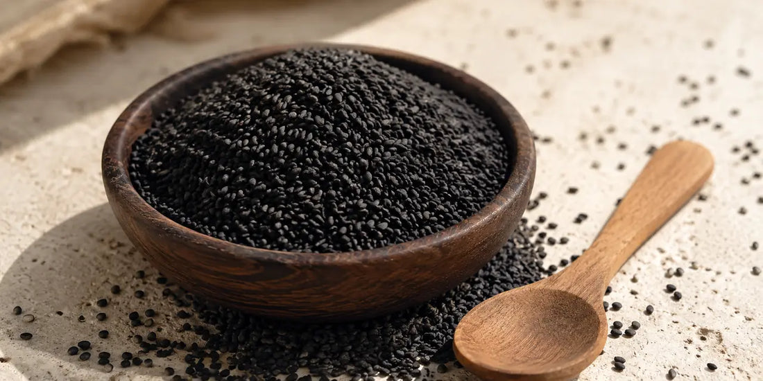 Shallow dark wooden bowl heaped with matte black sesame seeds and a plain wooden spoon on stone