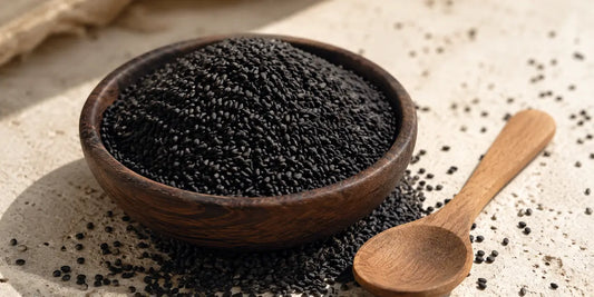 Shallow dark wooden bowl heaped with matte black sesame seeds and a plain wooden spoon on stone