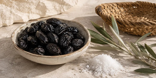 Ceramic bowl of wrinkled black table olives beside coarse salt, a woven basket and olive leaves