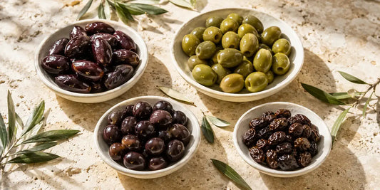Four white dishes of different cured olives on pale limestone with scattered olive leaves in warm daylight