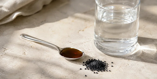 Small steel teaspoon of deep bronze oil beside a plain glass of water and scattered black seeds