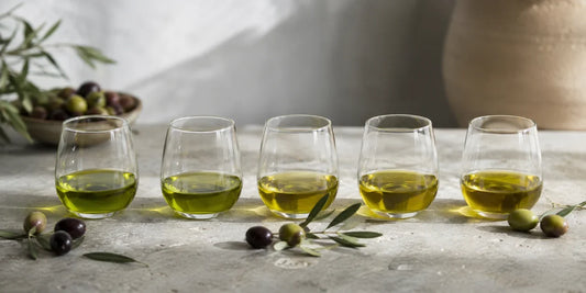 Five unbranded glass tasting cups of olive oil in varying green-gold shades beside fresh olives on pale stone