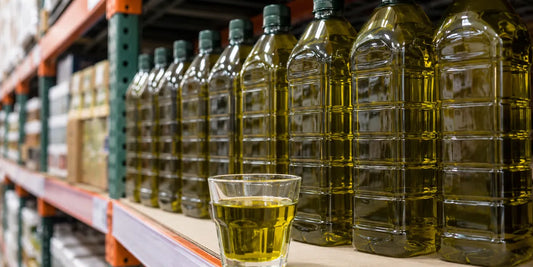 Large olive oil bottles lined up on a bright warehouse-style shelf with a golden-green oil glass in front in soft daylight