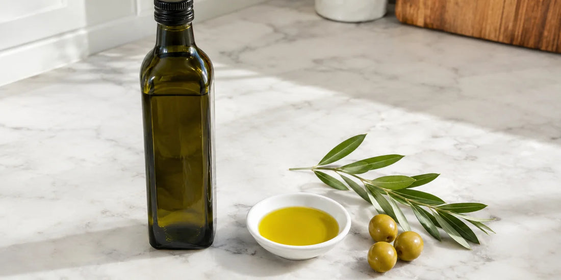 Dark glass oil bottle beside a white dish of rich golden-green olive oil and olives on pale marble