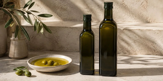 Two unlabelled dark green olive oil bottles beside a shallow dish of golden-green oil on pale limestone