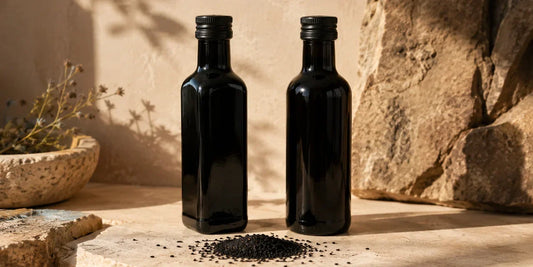 Two dark glass black seed oil bottles side by side on a pale stone surface with Nigella seeds in warm directional light