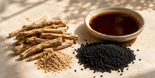 Dried ashwagandha roots beside matte black Nigella sativa seeds and a small dish of deep amber oil on pale stone