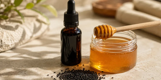 Dark glass black seed oil bottle beside a jar of golden manuka honey with a wooden dipper and scattered black seeds on pale stone