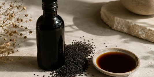 Dark glass black seed oil bottle beside scattered matte black nigella seeds and a dish of deep amber oil on pale stone