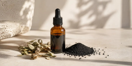 Unbranded amber glass dropper bottle of dark herbal tonic beside scattered matte black nigella seeds on pale stone