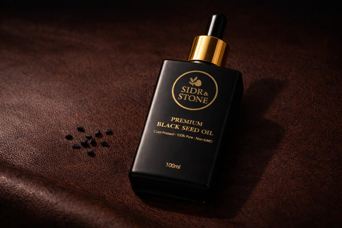 Matte black glass bottle of black seed oil with gold dropper resting on a dark leather surface