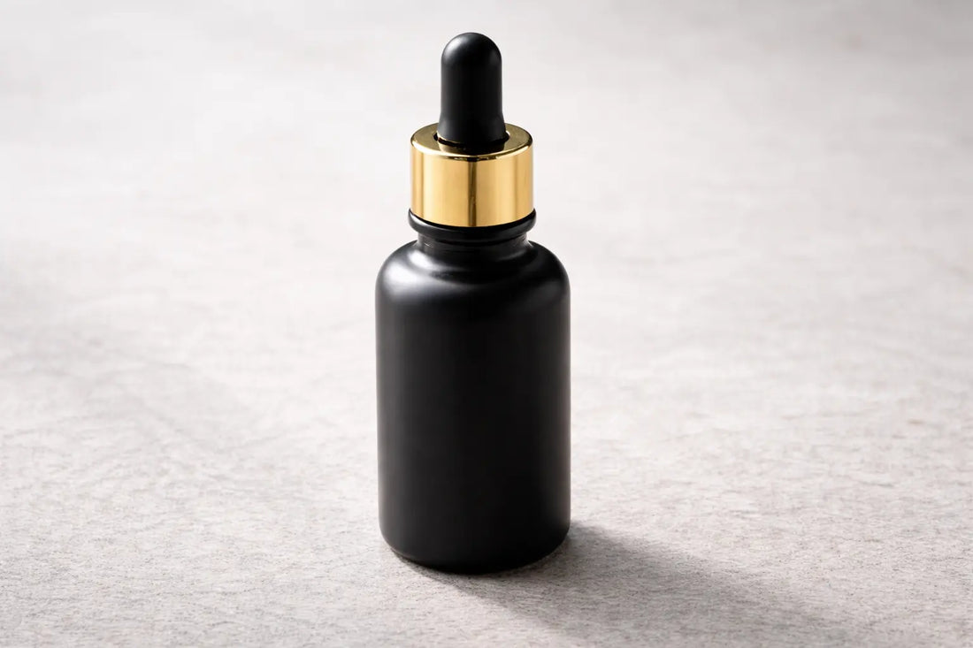 Matte black glass bottle of black seed oil with gold dropper on a neutral stone surface in calm lighting