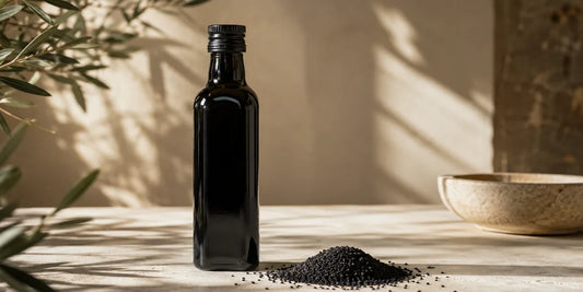 Dark glass black seed oil bottle beside a heap of matte black seeds on a pale stone surface in warm light