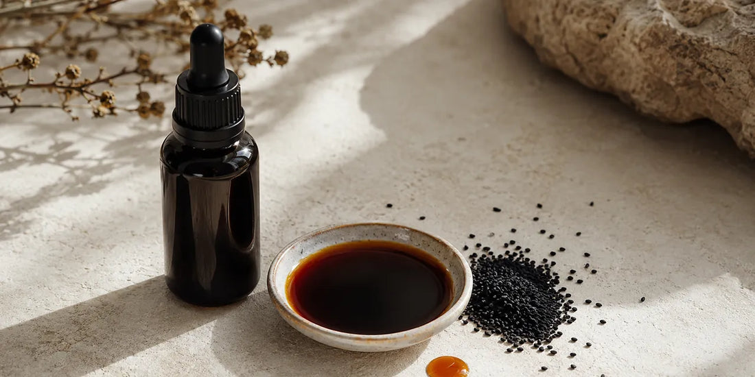 Dark glass dropper bottle beside a dish of deep amber black seed oil and scattered matte black seeds on pale stone