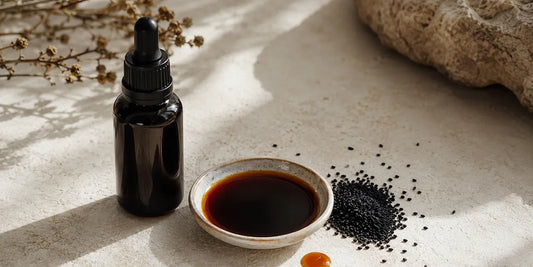 Dark glass dropper bottle beside a dish of deep amber black seed oil and scattered matte black seeds on pale stone
