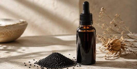Dark glass dropper bottle of deep amber black seed oil beside scattered black cumin seeds on pale stone in soft warm light