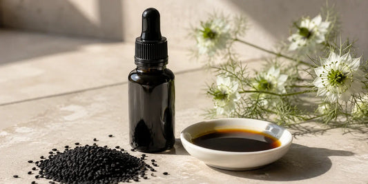 Dark glass black seed oil bottle beside scattered matte black cumin seeds and a dish of deep amber oil on pale stone