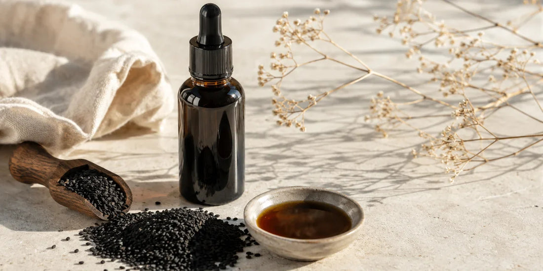 Dark glass black seed oil bottle beside scattered matte black cumin seeds and a dish of deep amber oil on pale stone