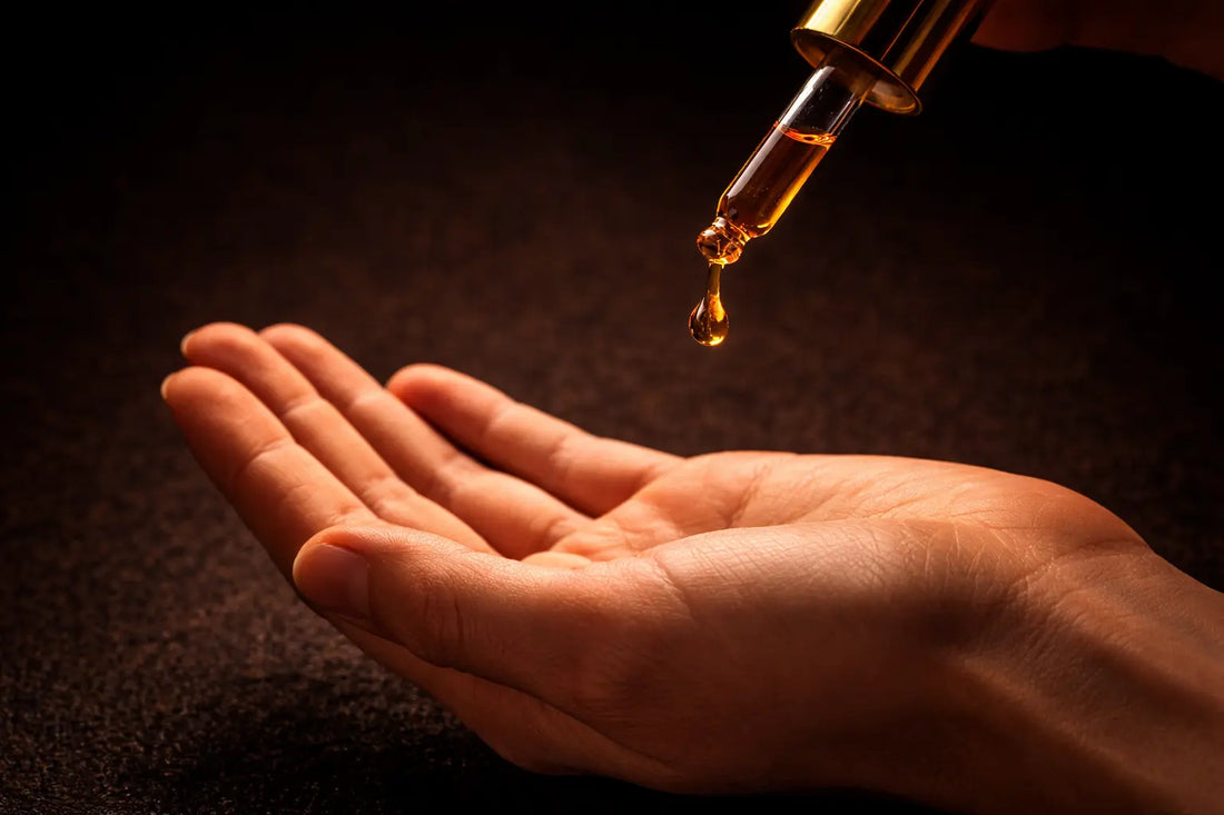 Glass dropper releasing a drop of dark bronze-amber black seed oil onto the back of a hand