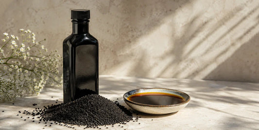 Dark glass black seed oil bottle beside scattered black seeds and a dish of deep amber oil on pale stone in calm light