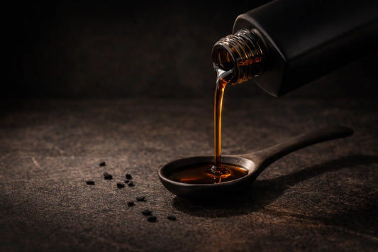 Dark bronze-amber black seed oil being poured into a ceramic teaspoon held over a dark stone surface