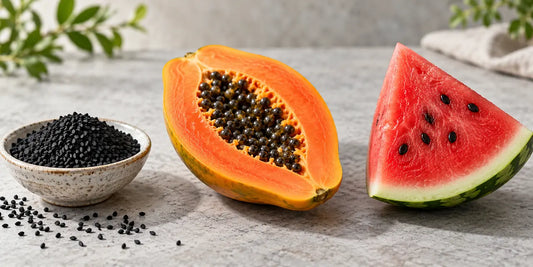 A dish of matte black nigella seeds beside a halved papaya with its seeds and a slice of seeded watermelon
