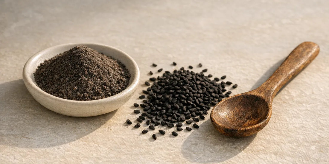 Pale dish of dark ground black seed powder beside scattered matte black seeds and a worn wooden spoon