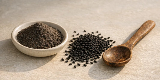 Pale dish of dark ground black seed powder beside scattered matte black seeds and a worn wooden spoon