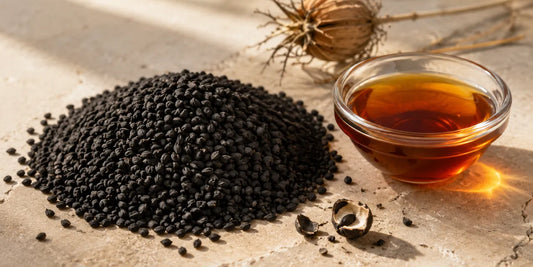 Matte black Nigella sativa seeds heaped beside a small glass dish of deep amber oil on pale stone