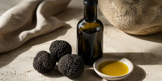 Three whole black truffles beside an unlabelled dark glass oil bottle and a dish of green-gold oil on pale stone