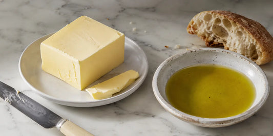 Block of butter on a white plate beside a dish of golden-green olive oil, knife and rustic bread